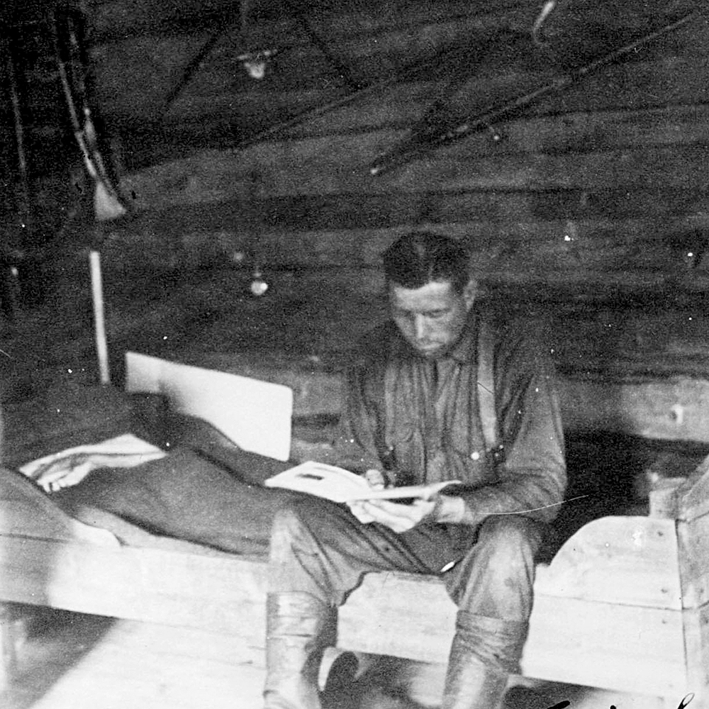 Kultamuseon kokoelmasta valokuva, jossa kullankaivaja Charles Hill istuu hirsikämpässä sängyllä lukemassa kirjaa.
